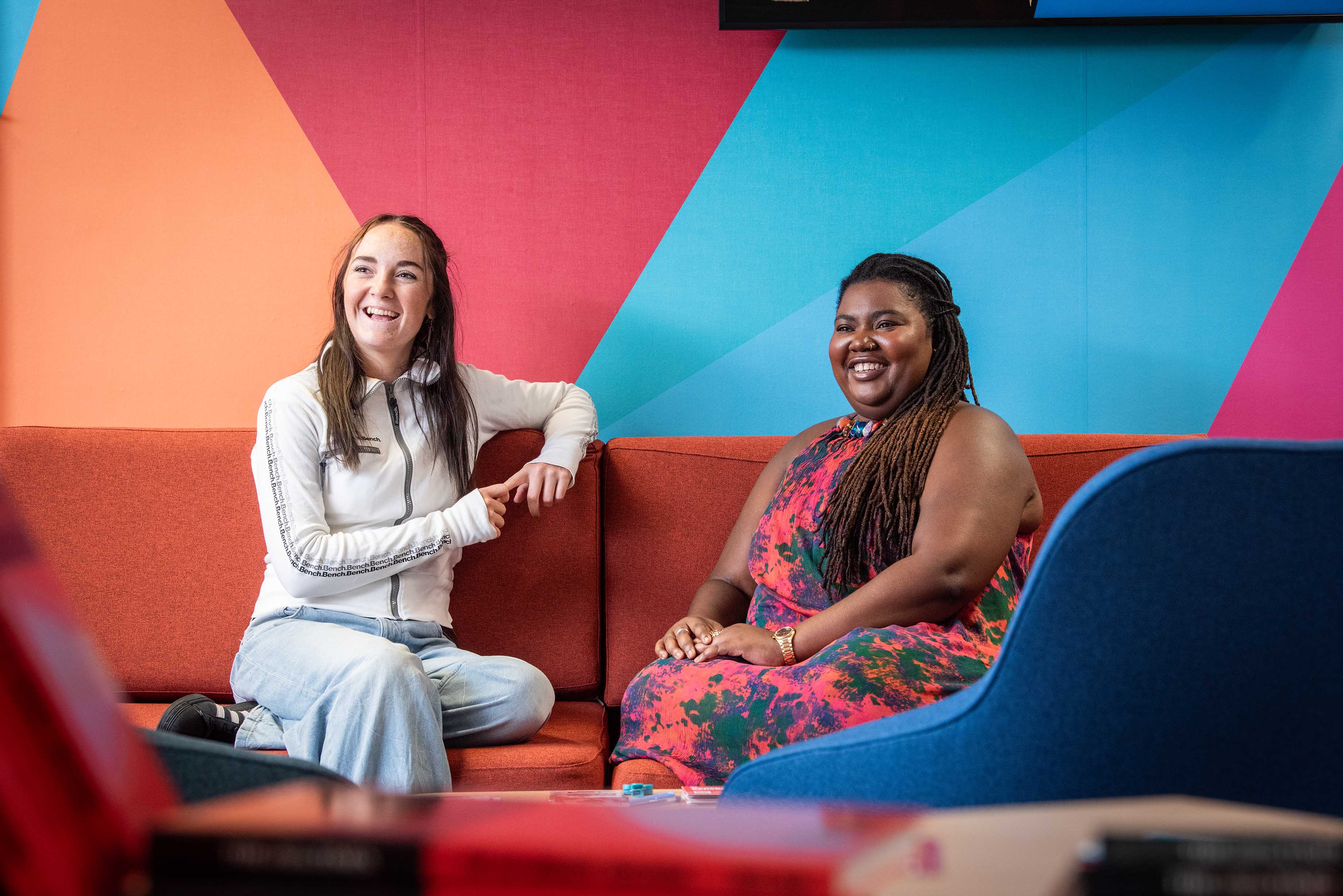 Amber and Sarauniya sit together on a sofa and smile at the camera against the backdrop of a colourful mural