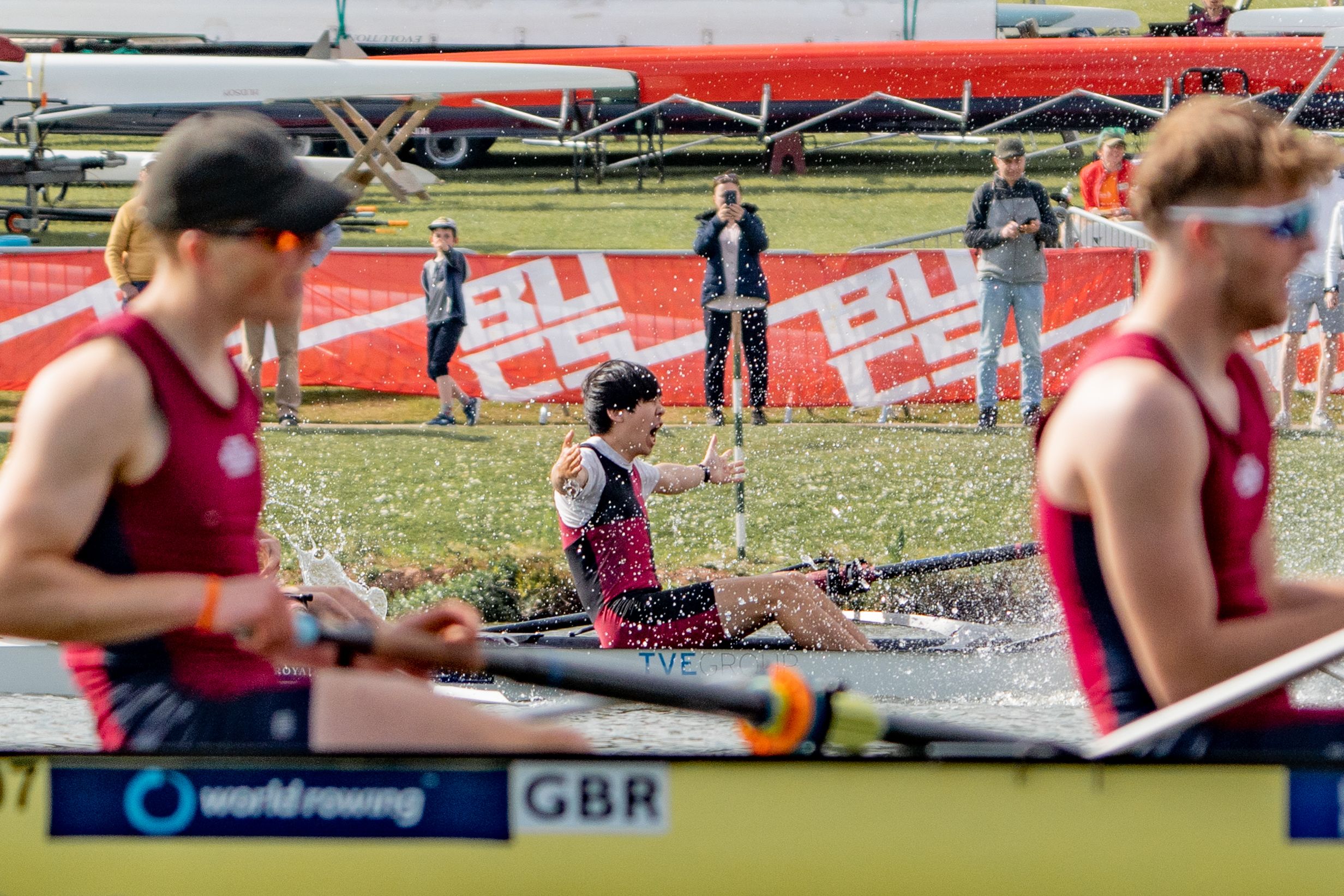 Ko is celebrating a win in his boat with his arms out wide, and cheering. Splashes of water surround him. Another rowing crew is in the foreground.