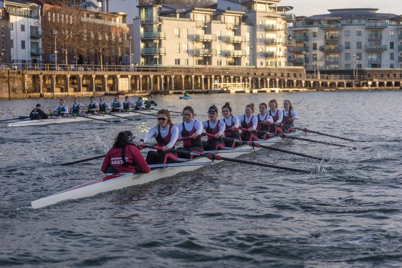 The UBBC Senior Women’s eight team competing against the City of Bristol Rowing Club on the harbour. The crew are wearing their white, black and red University of Bristol kit, and are mid-stroke. The sun is shining on the buildings lining the harbour in the background.