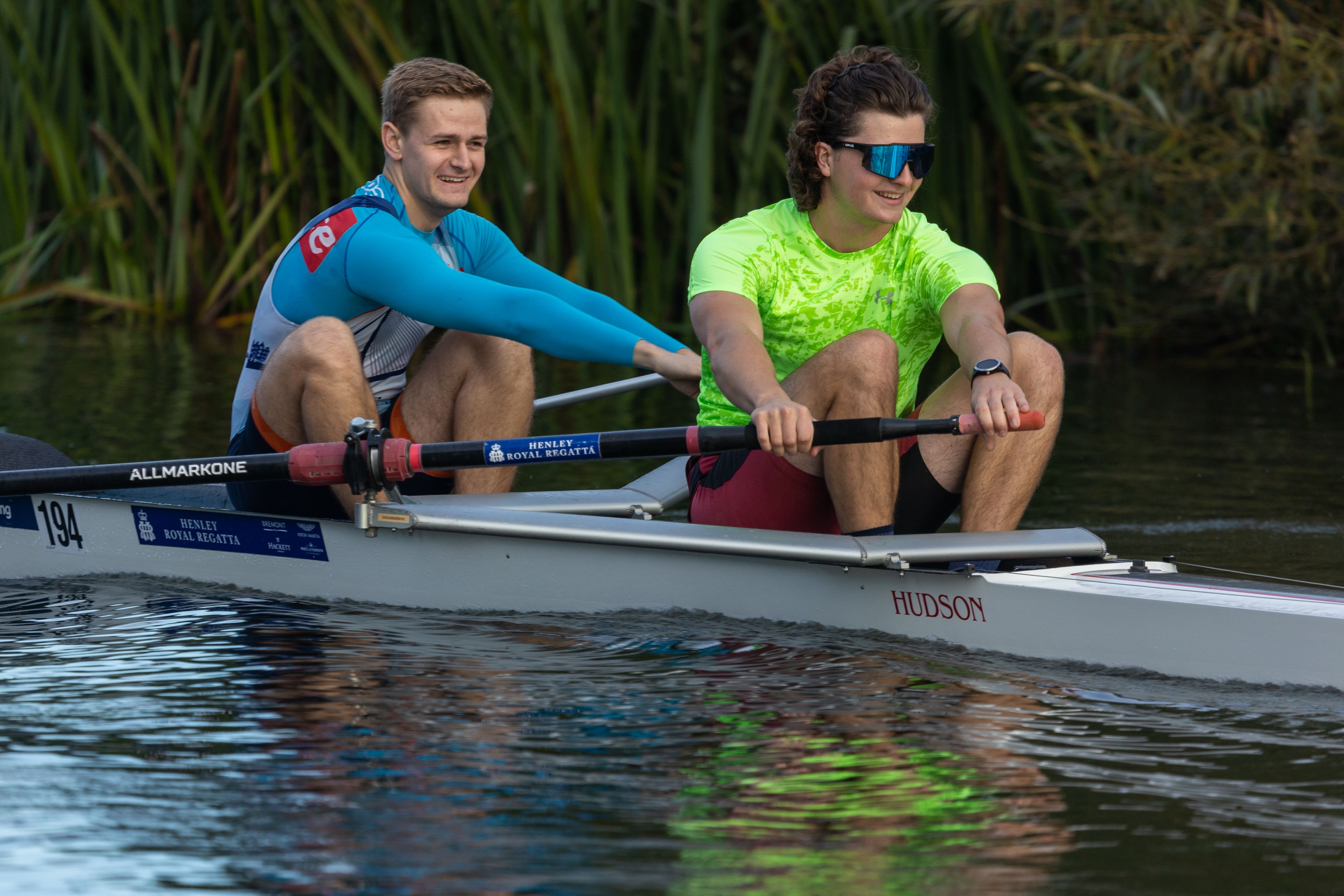 Tom Kuijlaars and Ben Crofts training on the water at Saltford. Both men are smiling as they row. Their boat is on the water in front of some tall reeds.