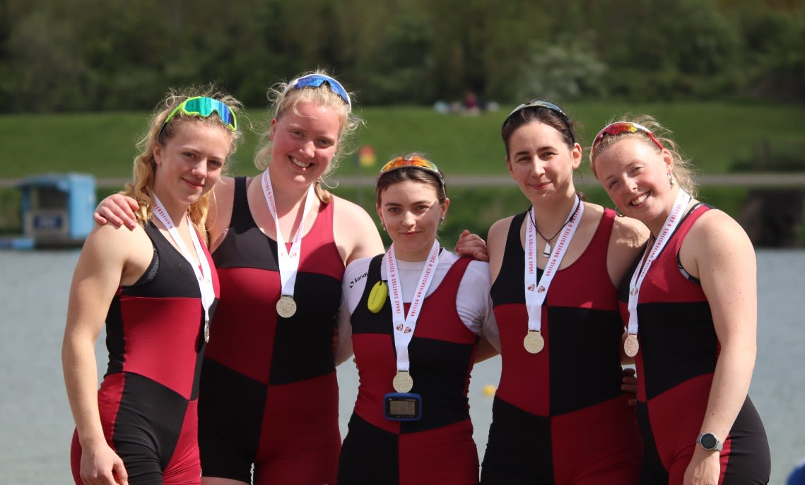 The UBBC Women’s coxed four crew are standing by the water’s edge with their arms around each other, smiling directly at the camera. They are all wearing their black and red University of Bristol rowing kit, and have silver medals around their necks.