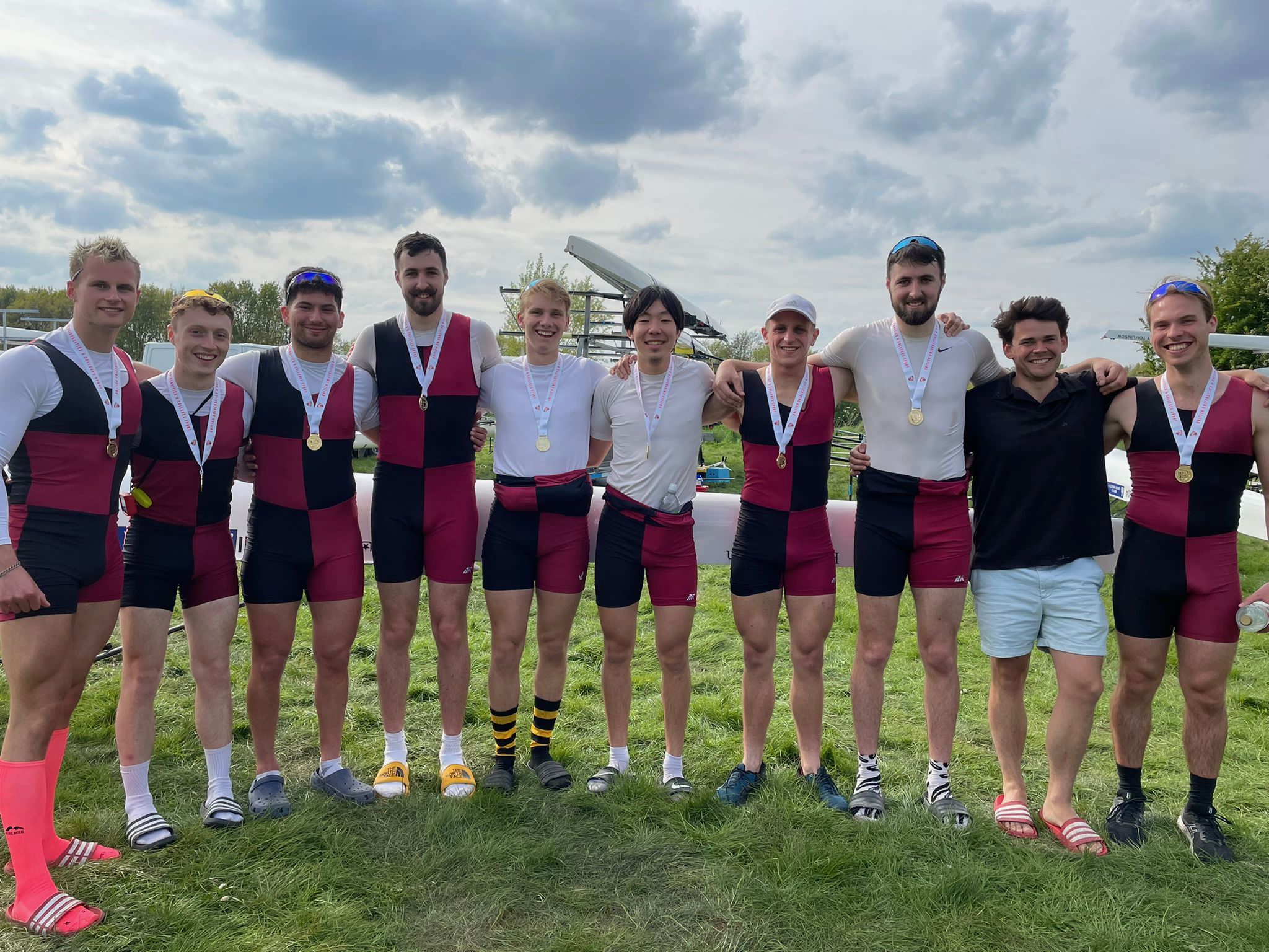The UBBC Senior Men’s coxed eight crew are standing in a line with their arms around each other on a grassy area near their boat. They are all smiling at the camera, and are wearing their white, black and red University of Bristol rowing kit. They have gold medals around their necks.