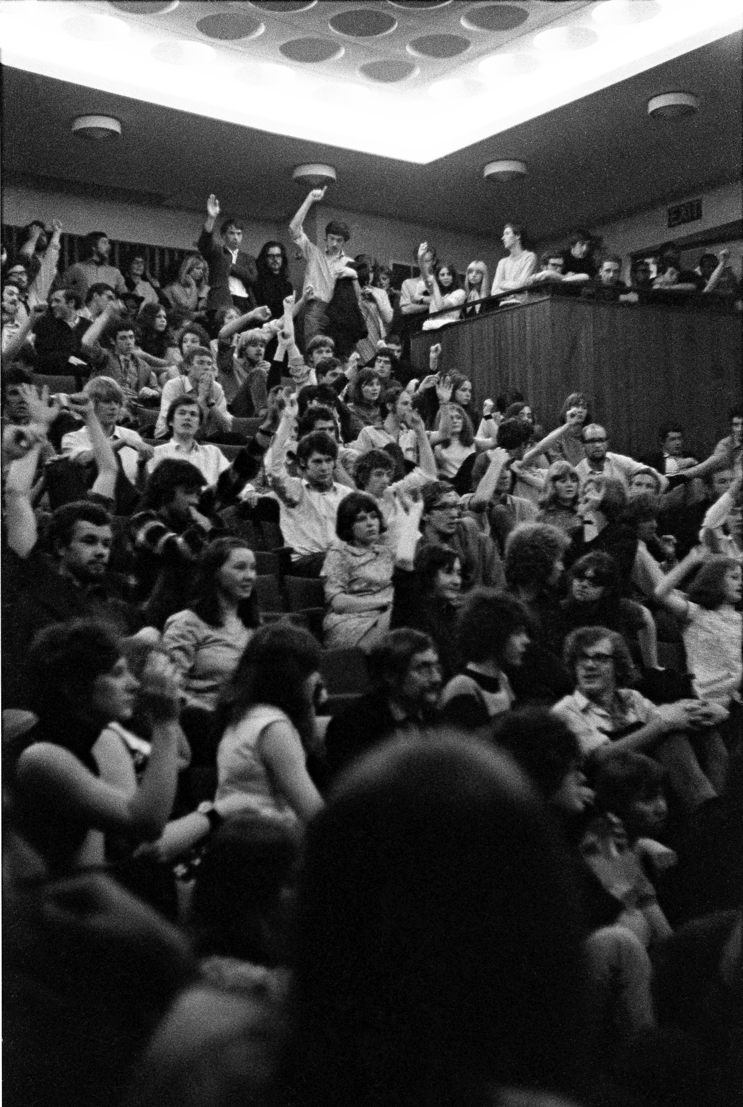 Around 70 students, some sitting and some standing, in a lecture theatre. Many have their hands raised, as part of a voting process.