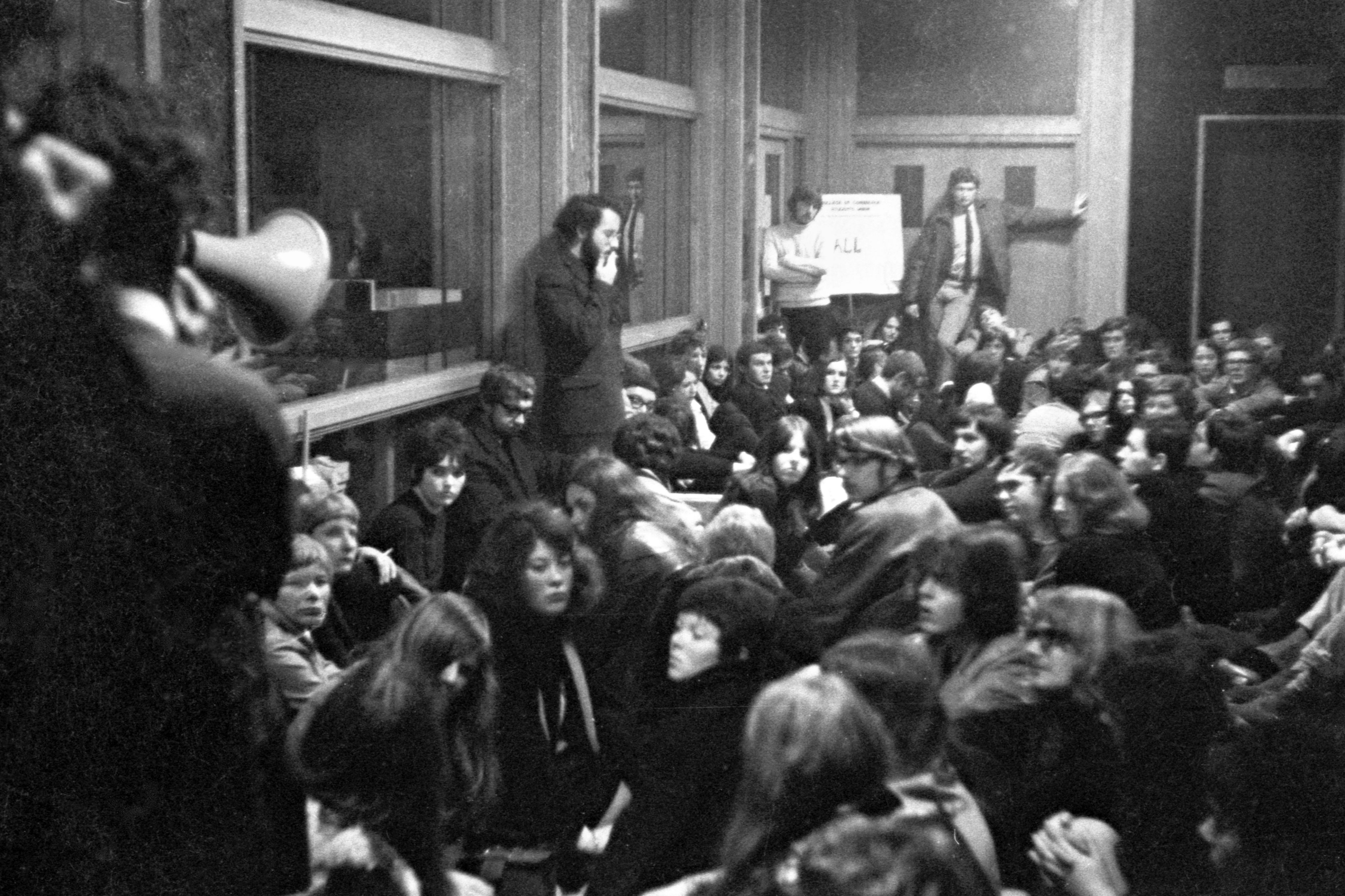 Around 30 students are standing close together in the same room. Four students are standing on a raised ledge, higher than their peers, and one is holding a megaphone.
