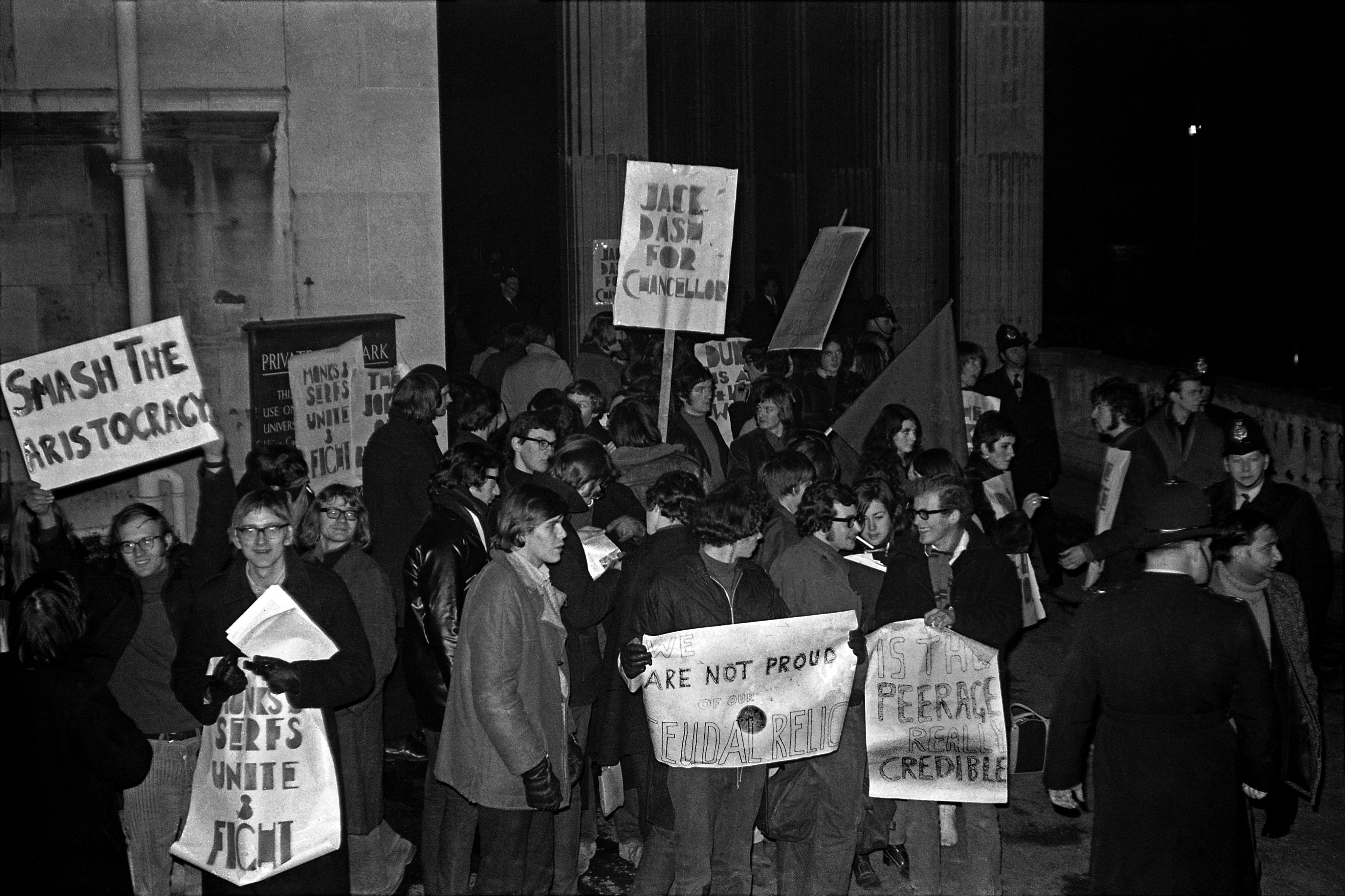 A group of around twenty students stand outside the Victoria Rooms, a University of Bristol building. Some hold placards, including one that reads 'Smash the Aristocracy' and another that reads 'Jack Dash for Chancellor'.